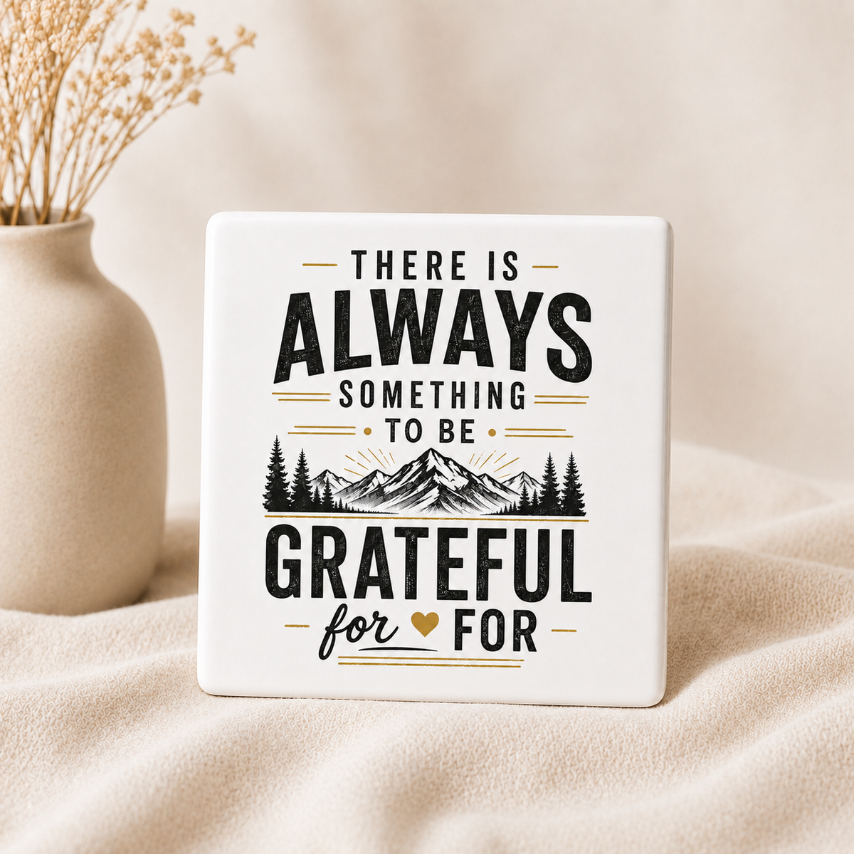 A square white ceramic coaster or sign with a black and gold quote that reads 'THERE IS ALWAYS SOMETHING TO BE GRATEFUL FOR' with a mountain and pine tree graphic, standing on beige fabric next to a small vase with dried stems.