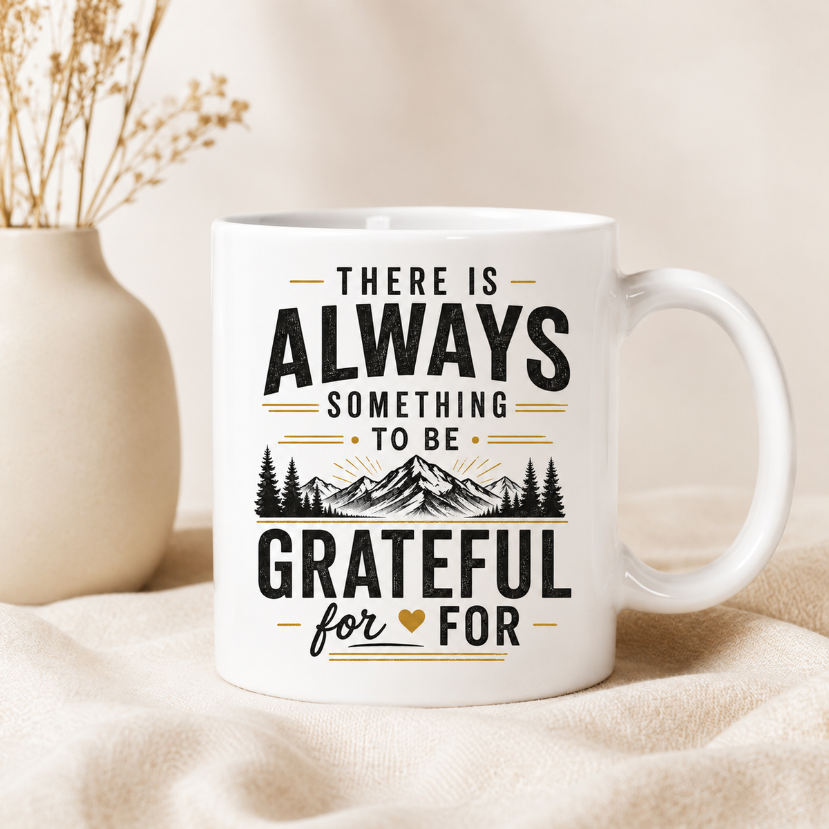 A white ceramic mug with a black and gold inspirational quote reading 'THERE IS ALWAYS SOMETHING TO BE GRATEFUL FOR' with a mountain and pine tree graphic, styled on a beige fabric surface next to a small vase with dried stems.