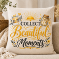 A square throw pillow in a warm cream fabric, printed with the phrase 'COLLECT Beautiful Moments' in black and gold lettering, surrounded by decorative illustrations of a sun, a mason jar, flowers, and a photo frame. The pillow is styled on a beige sofa with a knit blanket and dried grass decor in the background.