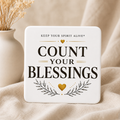 A square white decorative plaque with rounded corners featuring the printed phrase “COUNT YOUR BLESSINGS” in large black letters, with gold heart and leaf accents. The plaque is styled on beige fabric next to a small white vase with dried stems.