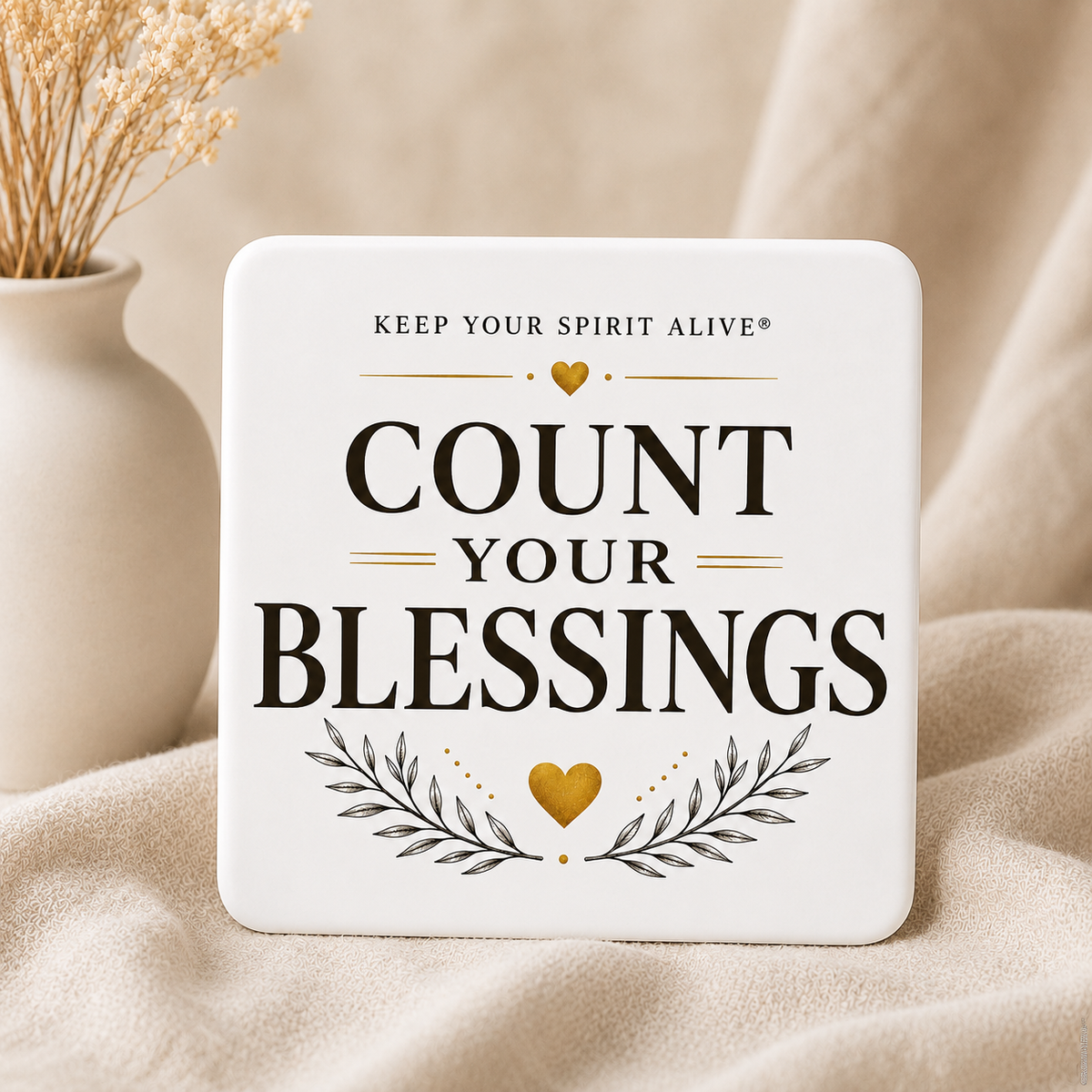 A square white decorative plaque with rounded corners featuring the printed phrase “COUNT YOUR BLESSINGS” in large black letters, with gold heart and leaf accents. The plaque is styled on beige fabric next to a small white vase with dried stems.