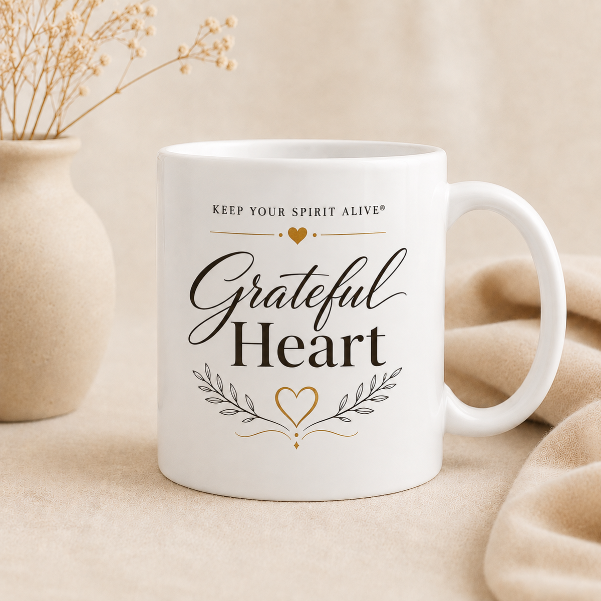 A white ceramic mug with the words 'Grateful Heart' and 'KEEP YOUR SPIRIT ALIVE' printed on it, styled on a beige fabric surface with a small vase of dried stems in the background.