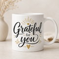 A white ceramic coffee mug with the phrase 'Grateful for you' in black script and gold decorative accents, set against a warm beige background with a blurred vase of dried flowers.