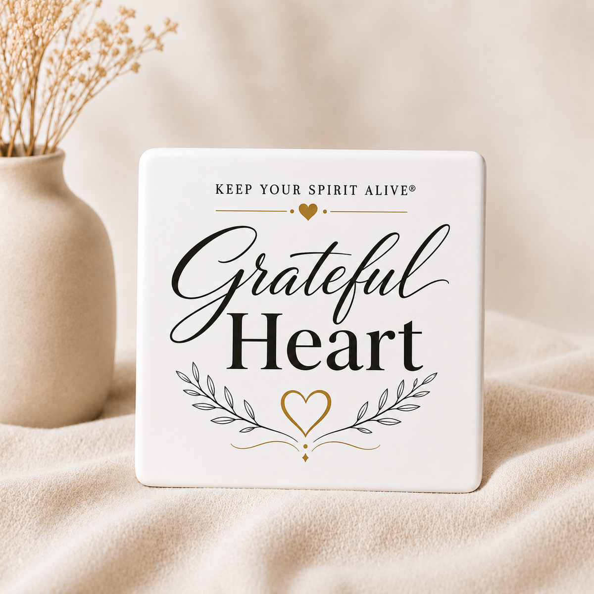 A square white ceramic coaster with the words 'Grateful Heart' in black script and serif fonts, plus a gold heart and leaf design, displayed on a beige fabric surface next to a vase with dried flowers.
