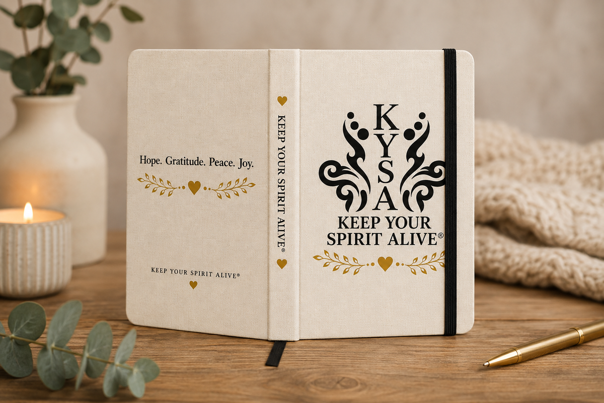 An open cream-colored notebook or journal on a wooden table, featuring the words 'Keep Your Spirit Alive' in black and gold text with decorative flourishes. The scene is styled with a candle, eucalyptus leaves, a knit blanket, and a gold pen in a warm indoor setting.