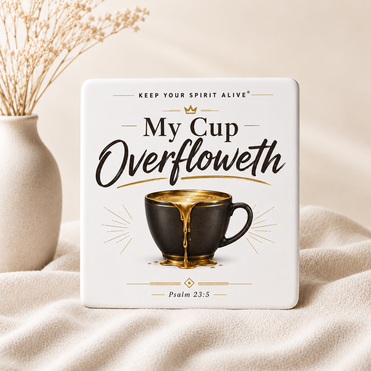 A square white ceramic coaster or small plaque with printed text reading 'My Cup Overfloweth' and an illustration of a dark cup with gold liquid dripping out, styled on beige fabric beside a vase with dried stems.