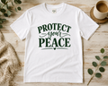 A white short-sleeve graphic T-shirt laid flat on a light wood surface, with dark green text reading “PROTECT your PEACE” and a small heart detail. The shirt is styled with eucalyptus leaves, a woven blanket, and a cup in the background.