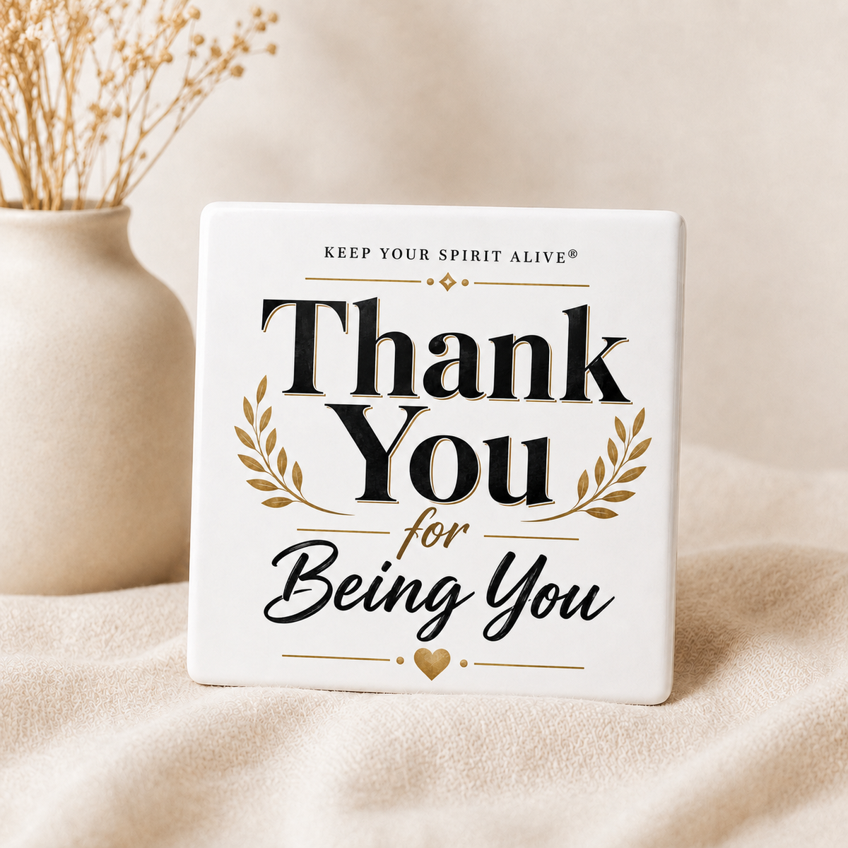 A square white ceramic sign with the printed message 'Thank You for Being You' in black and gold lettering, styled on a beige fabric surface next to a small vase with dried stems.