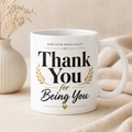 A white ceramic mug with a gold and black 'Thank You for Being You' graphic, styled on a beige fabric surface next to a small vase with dried flowers.