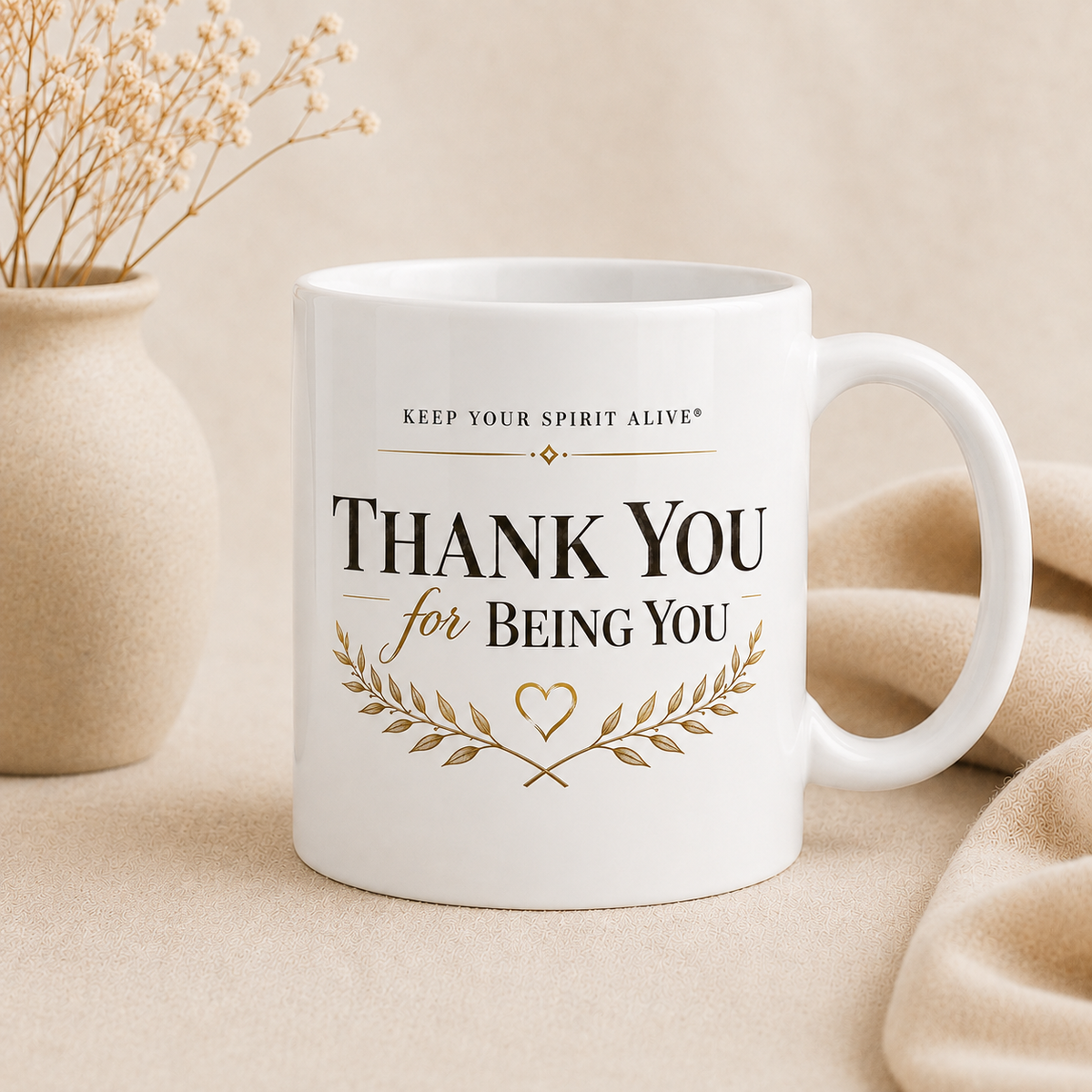 A white ceramic mug with the printed message 'THANK YOU for BEING YOU' in black and gold lettering, styled on a beige fabric surface with a beige vase of dried stems in the background.